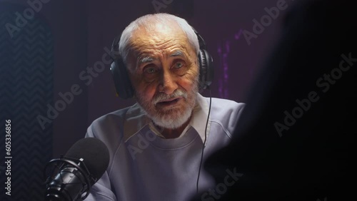 The old man creates a podcast in a professional studio. Senior male does online streaming interviews. Elderly man blogger speaking in microphone or make vlog. Recording audio content for social media photo