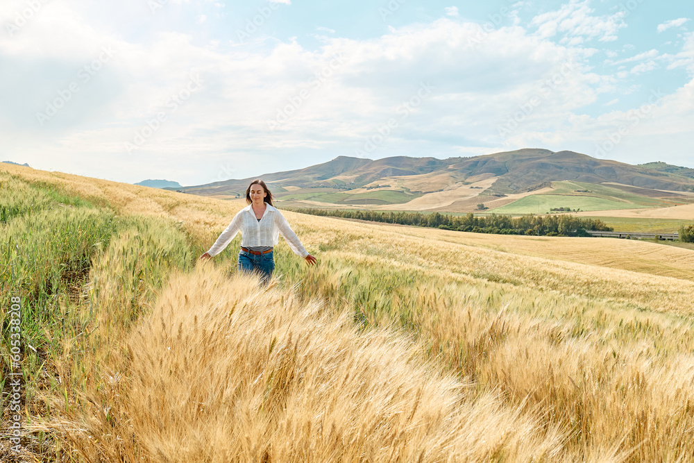 Woman walking in golden wheat field in hot summer sun and blue sky with white clouds with mountains hill landscape in background.