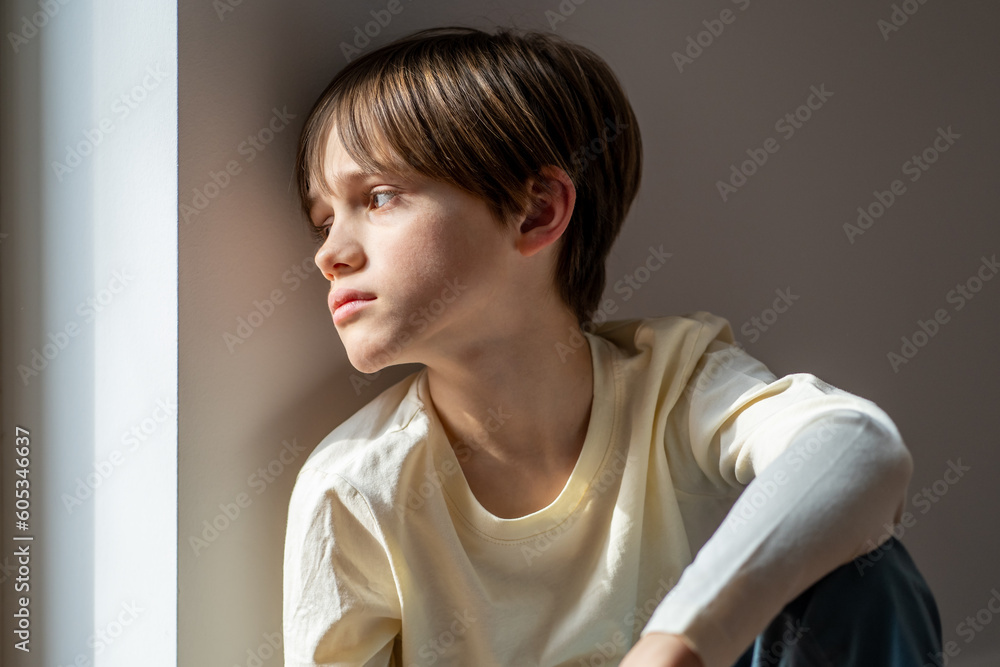 Loneliness in children. Sad boy child sitting on windowsill at home, looking out window. Orphan kid feeling lonely or isolated, waiting for parents in orphanage - obrazy, fototapety, plakaty 