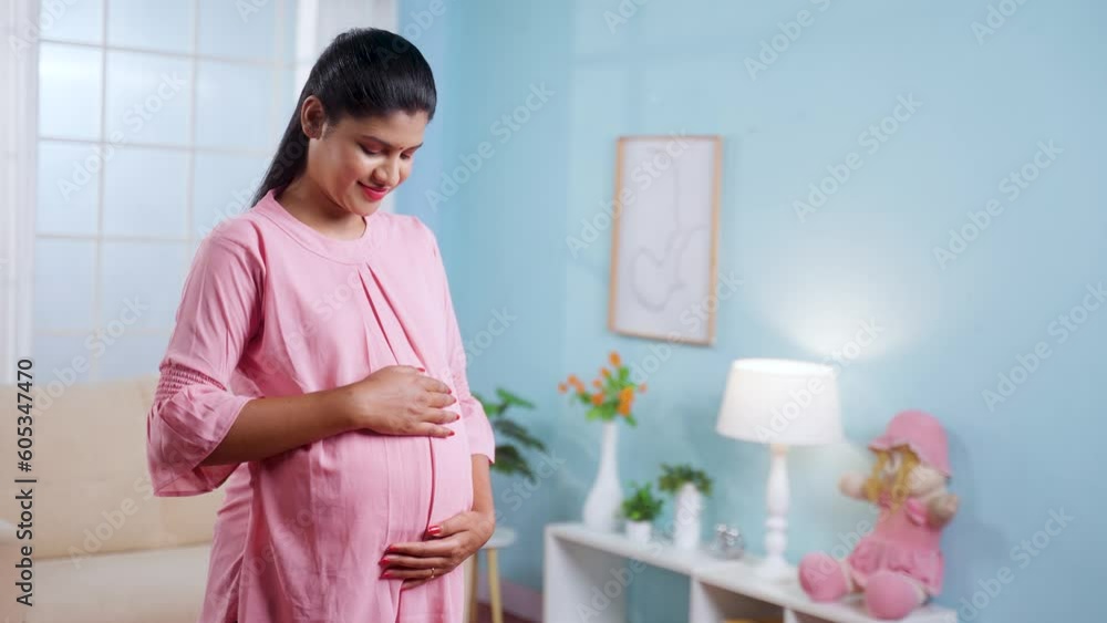 Happy smiling Indian Pregnant woman standing by feeling tummy while looking camera at home - concept of motherhood, expecting baby and parenthood Happy smiling Indian Pregnant woman standing by feeling tummy while looking camera at home - concept of motherhood, expecting baby and parenthood