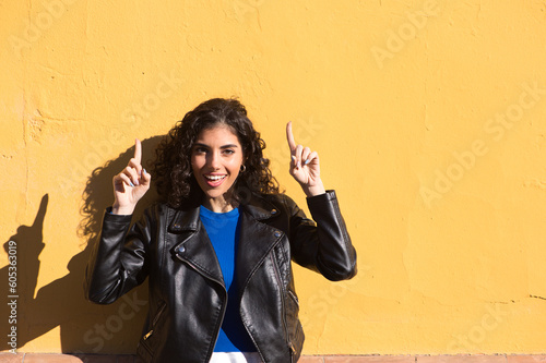 Young, beautiful Hispanic woman in casual clothes points with her index fingers up while smiling self-confident and happy. Background yellow wall