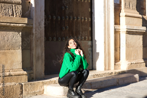 pretty young brunette woman with curly hair and green woollen coat is sitting on a step at the entrance of a building in seville, spain. The woman is happy and enjoying her holiday in the city.