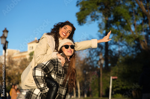 two young, beautiful Spanish women who are friends have fun playing on top of each other while they are walking and enjoying the sunny winter day. The women are wearing winter clothes.