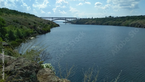 Bridge over the river. Khortytsia island, Dnipro river before russians blew up Kakhovka Dam, Zaporizhzhia, Ukraine. Green hills, rocks, blue sky and clouds. Calm water. photo