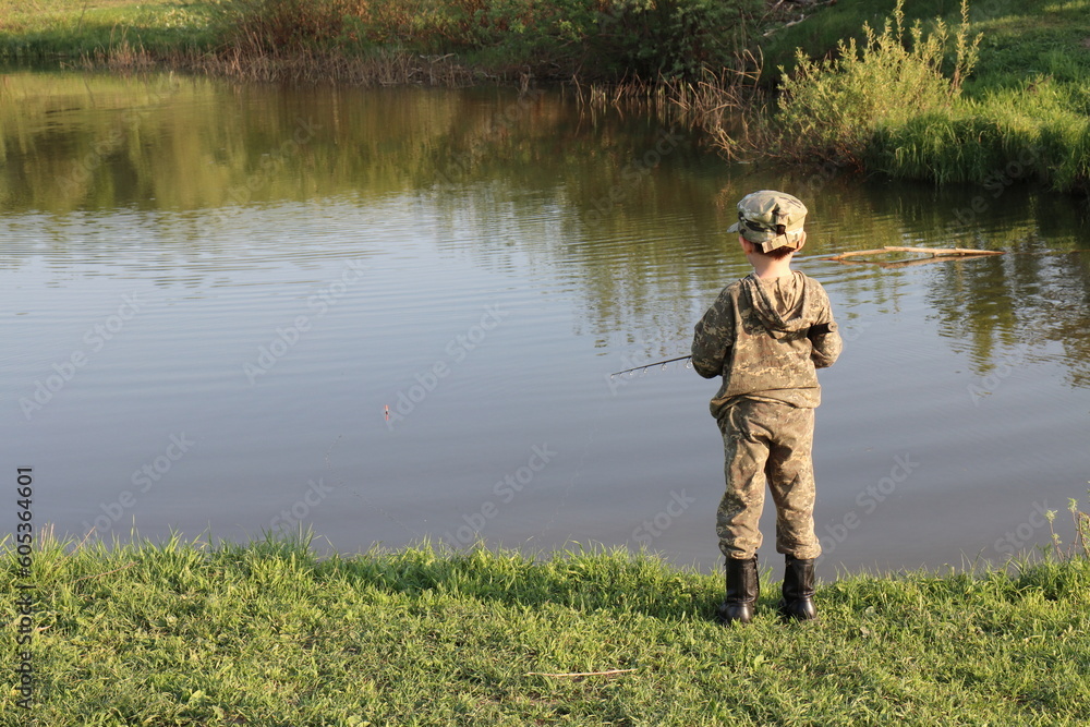 A boy in camouflage clothing and black boots is fishing with a fishing rod on the bank of a quiet river on a spring evening A boy in camouflage clothing and black boots is fishing with a fishing rod on the bank of a quiet river on a spring evening
