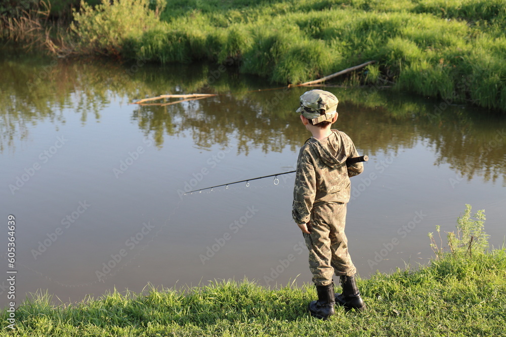 A boy in camouflage clothing and black boots is fishing with a fishing rod on the bank of a quiet river on a spring evening A boy in camouflage clothing and black boots is fishing with a fishing rod on the bank of a quiet river on a spring evening