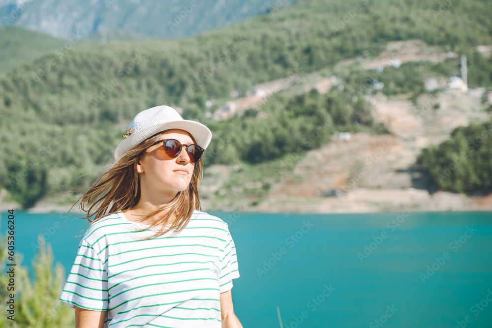 Fototapeta premium Smiling woman in hat and sunglasses with wild hair standing near mountains lake on background. Positive young woman traveling on blue lake outdoors travel adventure vacation.