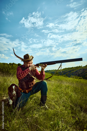 Portrait with male hunter wearing retro clothes with hat holding gun and shooting with his companion dog English springer spaniel over nature landscape background