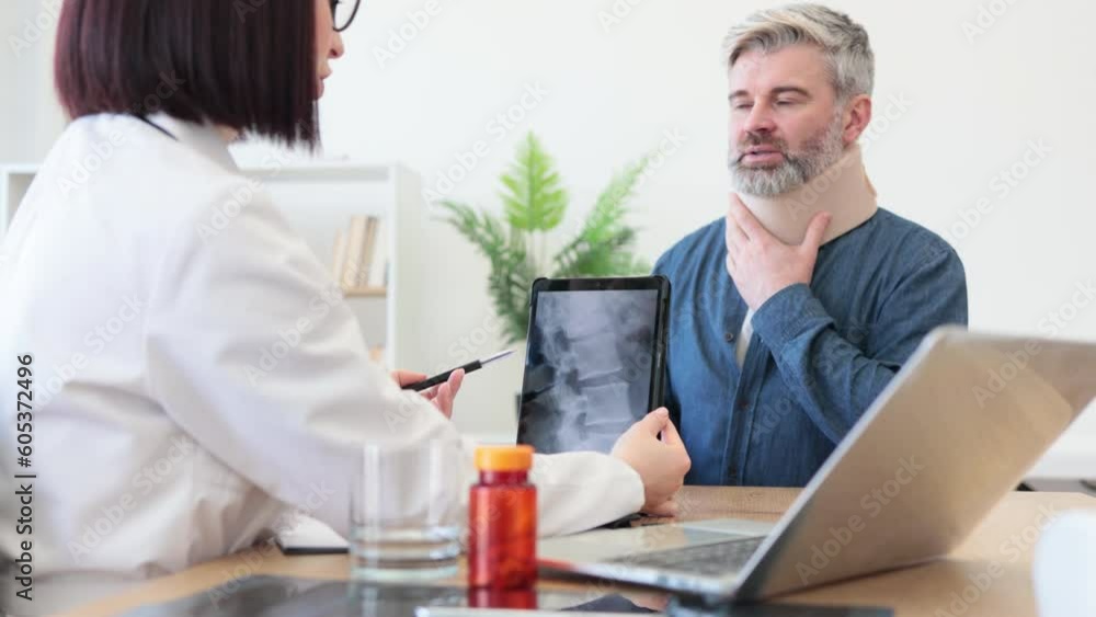 Back view of female doctor holding digital tablet while sitting in front of smiling caucasian man in cervical collar. Health professional consulting mature patient about neck injury using CT scan. Back view of female doctor holding digital tablet while sitting in front of smiling caucasian man in cervical collar. Health professional consulting mature patient about neck injury using CT scan.