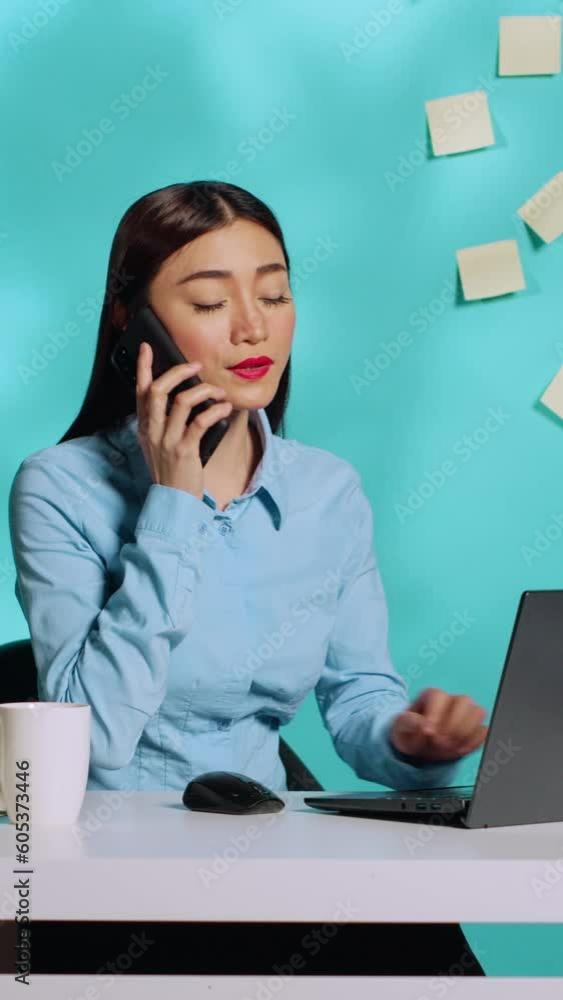 Smiling businesswoman receiving confusing information over phone call while working on laptop. Professional executive working at modern bright office desk over blue studio background