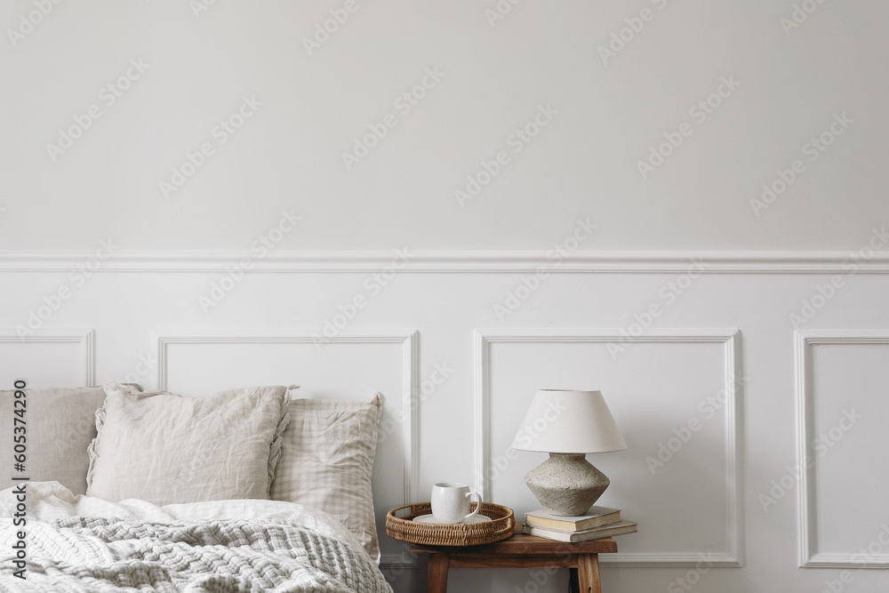 Cup of coffee on wicker tray. Textured clay table lamp, old books on vintage wooden bench, night stand. Bedroom view. Checkered beige pillows, linen blanket. Elegant Scandinavian interior, copy space. - obrazy, fototapety, plakaty 