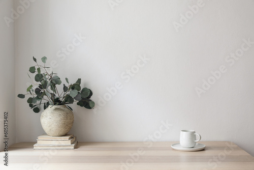 Minimal breakfast still life. Cup of coffee, tea. Empty white wall in sunlight table. Textured vase with silver eucalyptus leaves, branches on old books. Elegant Scandinavian working space, office