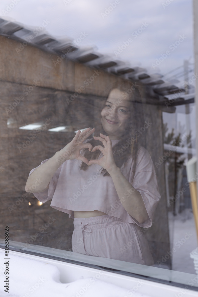 Fototapeta premium Photograph through glass. Young beautiful girl standing at the window and showing a heart