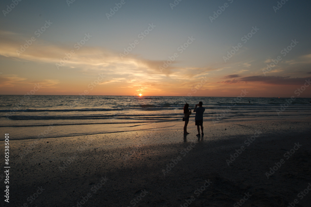 silhouette of man and woman  at Florida beach at sunset with blue sky and yellow colors with water and sand.. Taking pictures of sunset with cell phone . Sun on edge of horizon.