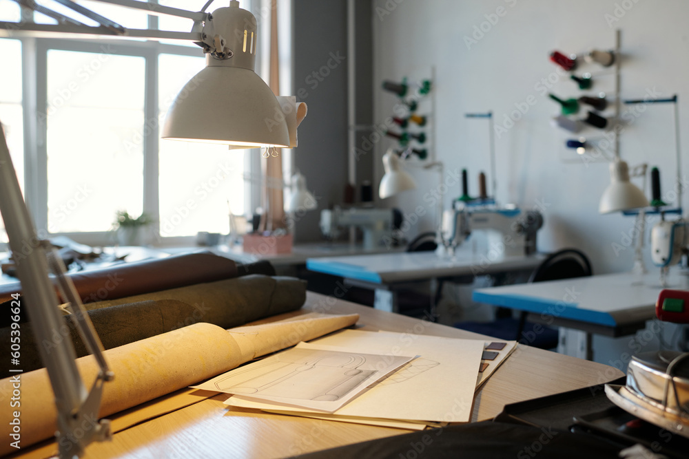 Focus on workplace of leatherworker with lamp, sketches and rolled leather textile of various colors against desks with sewing machines