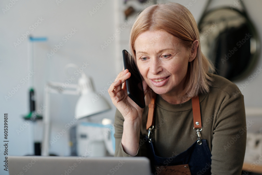 Smiling blond mature woman with smartphone by ear looking at laptop screen and talking to one of clients while specifying details of order