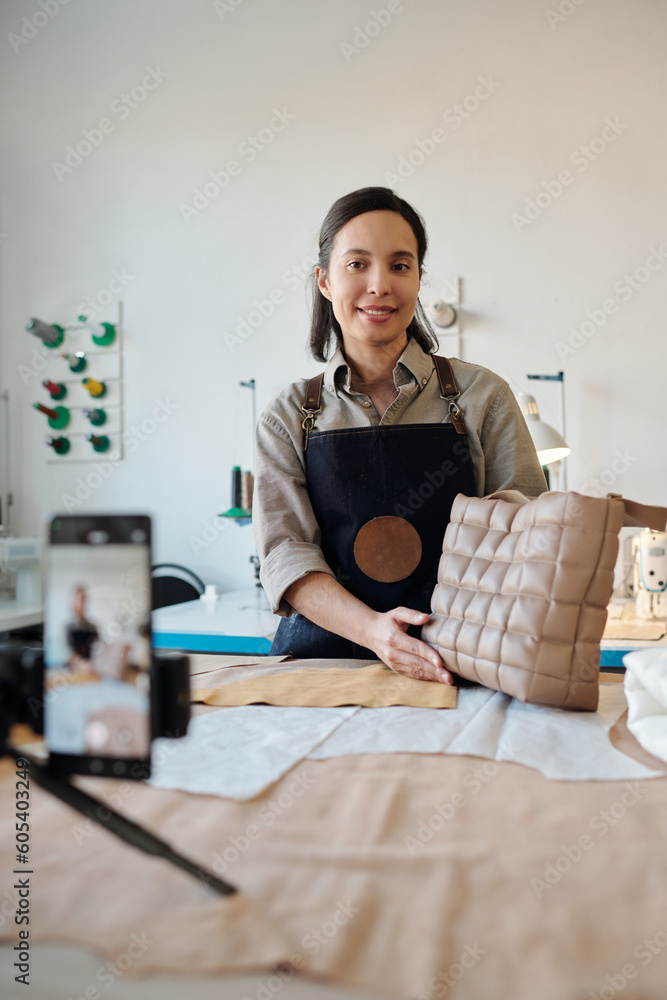 Young smiling creative woman in apron with new handmade leather bag looking at smartphone camera while giving masterclass in studio Young smiling creative woman in apron with new handmade leather bag looking at smartphone camera while giving masterclass in studio
