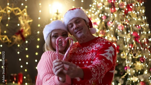 Guy and girl celebrate Christmas, New Year in santa hats hugging at each other and hold in hands Christmas candy cane in shape of heart against background of Christmas tree and lit New Year's garlands