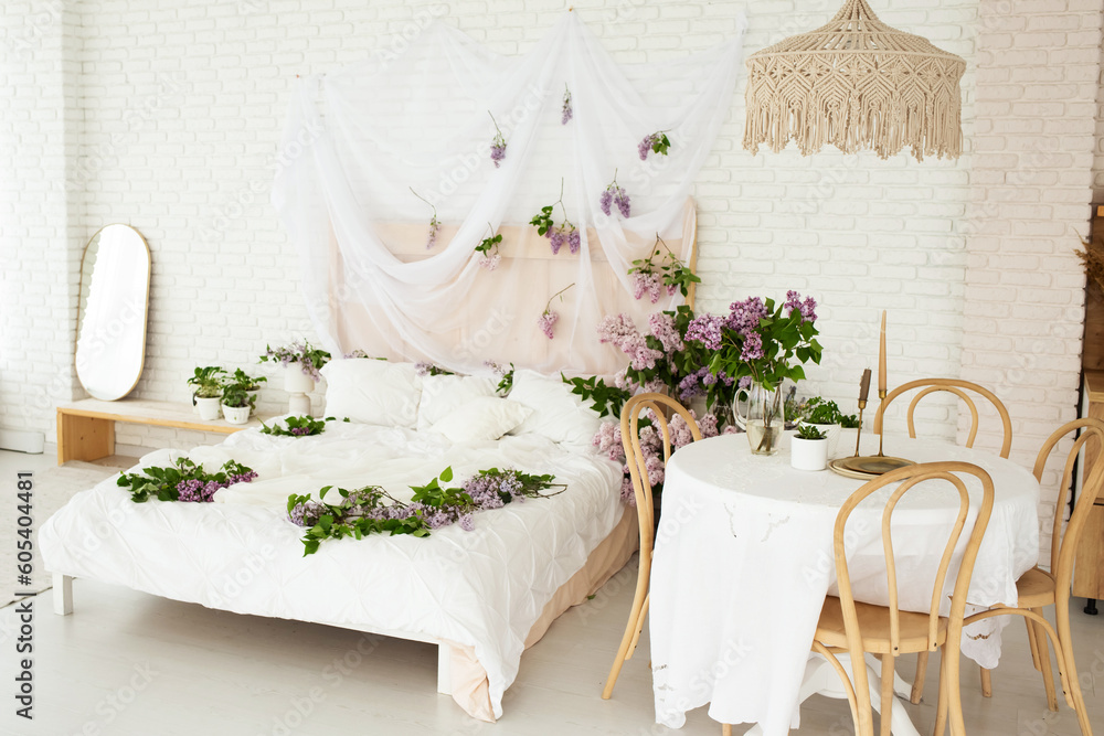 beautiful bright room in boho style with a wooden table and chairs and a large bed covered with a white bedspread and decorated with lilac flowers beautiful bright room in boho style with a wooden table and chairs and a large bed covered with a white bedspread and decorated with lilac flowers