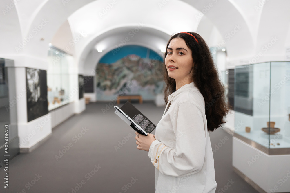Young Caucasian woman holds brochure and turn around visiting educational excursion in museum. Light hall in background. Concept of cultural exhibition Young Caucasian woman holds brochure and turn around visiting educational excursion in museum. Light hall in background. Concept of cultural exhibition
