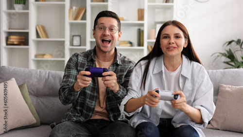 A boy and a girl are having fun playing video games with joysticks in the living room while sitting on the sofa. A couple in love on a date, enjoying the game, joking and having fun.