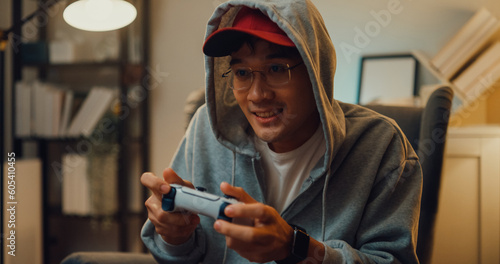 Happy young Asian man wear gray hoodie hold joystick controller excitement and focus while having fun playing video games sitting on the sofa in the living room in home at night.