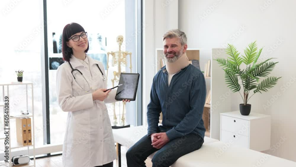 Cheerful young woman with digital tablet and smiling mature man in cervical collar posing in doctor's office. Therapist examining spine x-rays while happy patient sitting on exam couch indoors. Cheerful young woman with digital tablet and smiling mature man in cervical collar posing in doctor's office. Therapist examining spine x-rays while happy patient sitting on exam couch indoors.