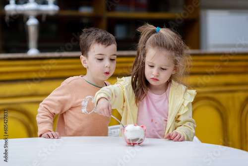 Cute little boys and a girl in yellow jackets together eat ice cream with two spoons from one bowl. Happy childhood