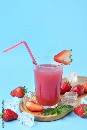 Board with glass of tasty strawberry juice and ice cubes on blue background