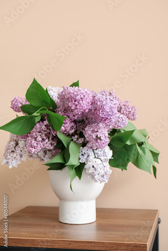 Vase with bouquet of lilac flowers on table near beige wall