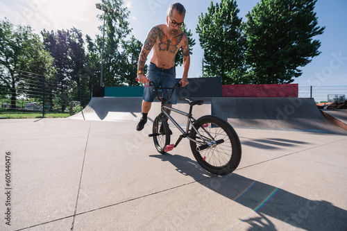 A focused mature shirtless urban man is performing stunts and tricks on his bmx in a skate park.
