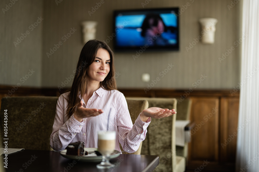 Young beautiful brunette woman sit in coffee shop cafe restaurant indoors points her hands to the side. Young beautiful brunette woman sit in coffee shop cafe restaurant indoors points her hands to the side.