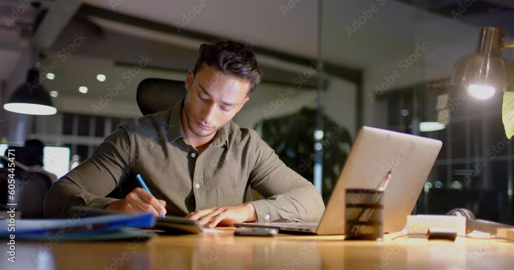 Busy biracial businessman at desk using laptop and making notes in office at night, slow motion Busy biracial businessman at desk using laptop and making notes in office at night, slow motion