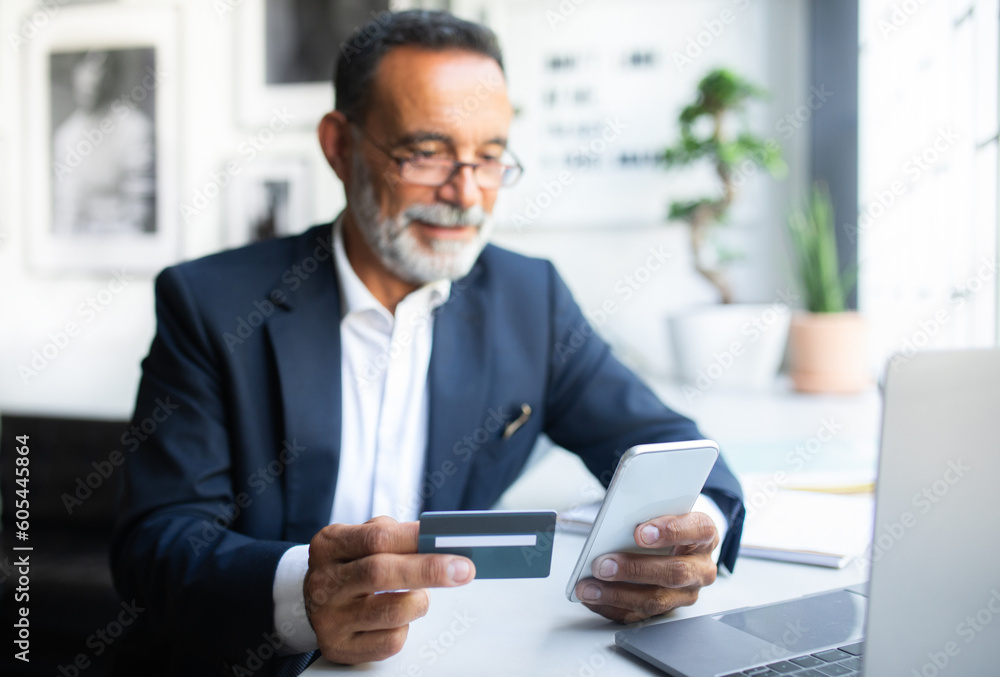 Smiling old caucasian man in suit and glasses with looks at phone, uses credit card at table in office Smiling old caucasian man in suit and glasses with looks at phone, uses credit card at table in office