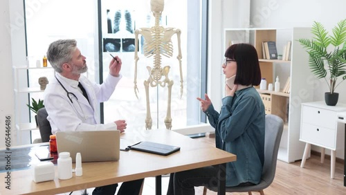 Side view of professional man in lab coat and attractive woman in C collar sitting at writing desk in doctor's office. Young caucasian lady attending outpatient appointment because of neck injury. photo