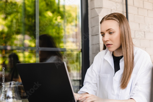 Confused young attractive blonde woman with casual hairstyle keeping hand on keyboard of laptop while looking perplexedly at screen with raised eyebrow sitting in cafe outside, serious girl working. photo