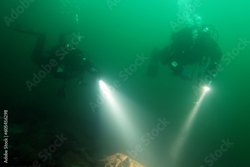 Two scuba divers pointing at an underwater stone formation with their torches