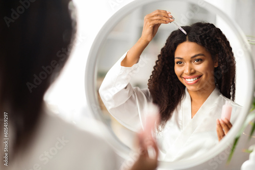 Slika na platnu Smiling Black Woman Applying Serum For Hair Repair While Standing Near Mirror