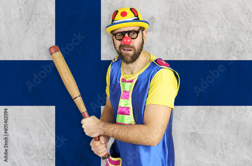 An insecure clown holds a bat in his hands against the background of the flag of Finland