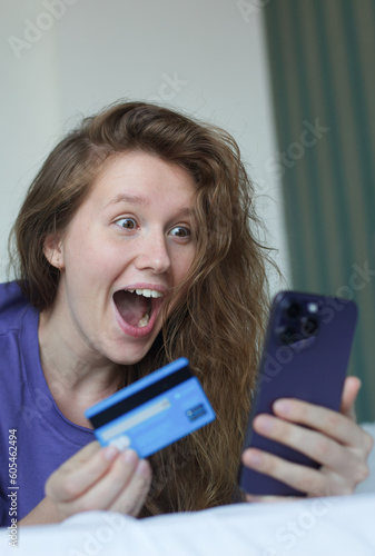 Portrait of happy excited amazed girl, shocked young successful joyful woman paying, holding credit bank card with phone, laptop celebrating victory, triumph, success at home. Online shopping
