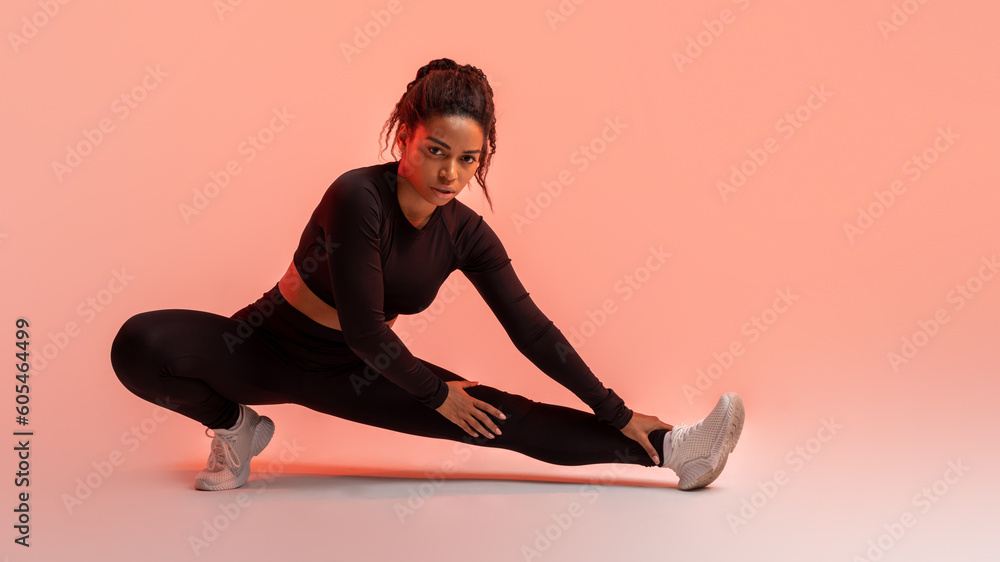 Studio perfection. Fit black lady mastering her exercise routine, stretching legs, peach neon background, panorama