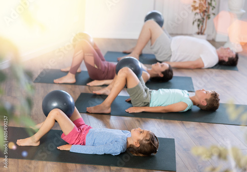 Sportive family members engaged in pilates lying on the floor on black mat holding softball between legs