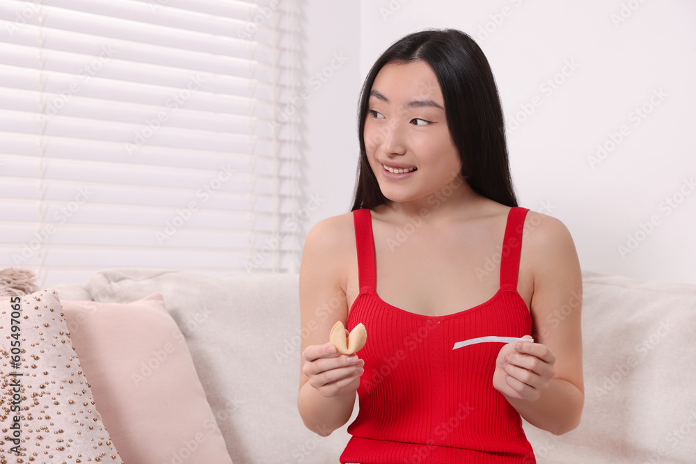 Asian woman holding tasty fortune cookie with prediction on sofa at home, space for text Asian woman holding tasty fortune cookie with prediction on sofa at home, space for text