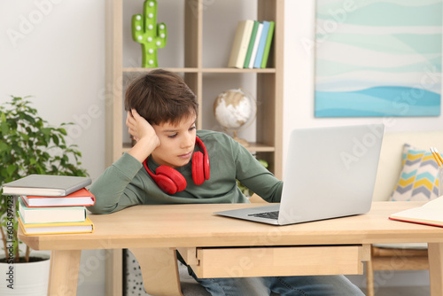 Tired boy with red headphones using laptop at desk in room. Home workplace © New Africa