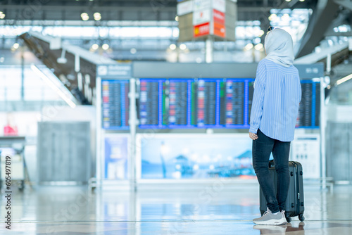 An Asian Muslim wearing a blue hijab is preparing for a vacation and she is at the airport, looking at the Flight Departure board.