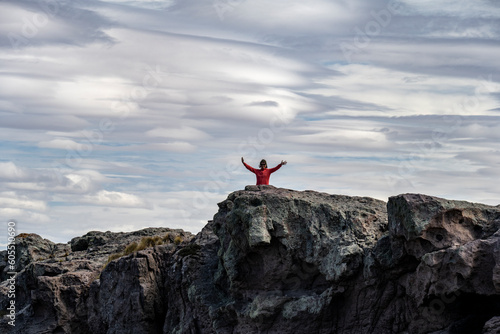 Third class woman with arms raised on a cliff with lenticular clouds in the background in Puerto Deseado, Santa Cruz, Argentina.