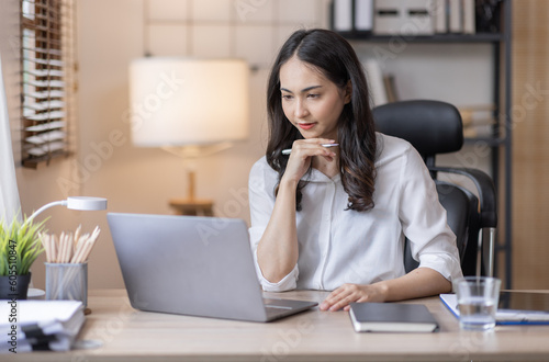 Business And Education Concept. Smiling young asian woman sitting at desk working on laptop writing letter in paper documents, free copy space. Happy millennial female studying using laptop