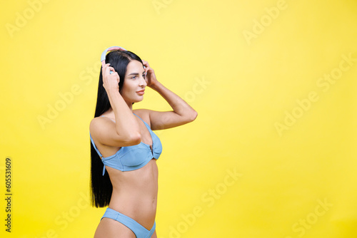Happy young woman wearing in blue swimsuit listening music in headphones and dancing, singing song and having fun isolated on yellow wall background studio. Summer rest concept