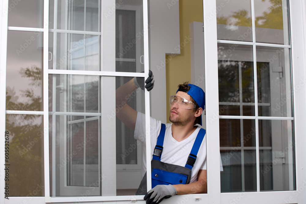 Pensive worker in blue jumpsuit and cap stands an open window holding protective net against insects with his hand. Young Caucasian builder performs installation of mask mesh on a plastic window
