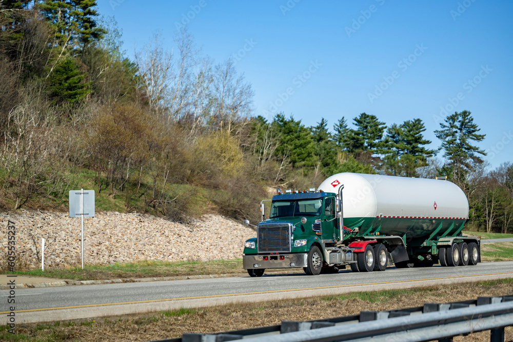 Day cab dark green big rig semi truck transporting flammable chemicals in a specialized durable tank semi trailer driving on the two lines highway road with rocks and green forest on the side